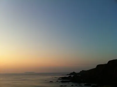 A coastal landscape at dusk showing calm ocean water, rocky shoreline and cliffs silhouetted on the right, a small island visible on the horizon, and a clear sky transitioning from orange near the horizon to deep blue above.
