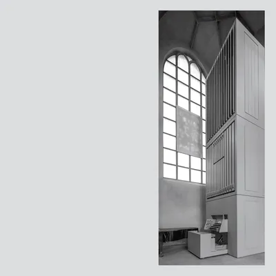 This is a black and white architectural photograph showing the interior of a church or chapel, featuring a large pipe organ mounted against a wall beside a tall arched window with grid-paned glass. The organ console with its keyboard and controls sits at the base of the tall wooden organ case.