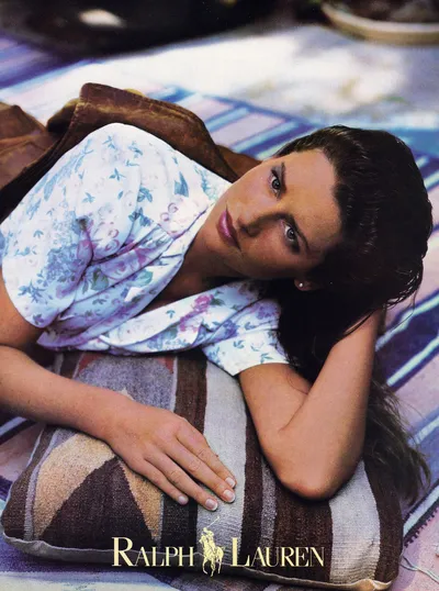 A vintage Ralph Lauren advertisement showing a woman with dark brown hair lying on a patterned blanket or rug, resting her head on her arm, wearing a white floral-print shirt and a brown jacket. The Ralph Lauren logo appears at the bottom of the image.