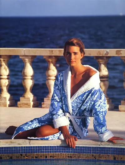 A fashion advertisement photo showing a woman with brown hair sitting beside a pool, wearing a blue and white floral bathrobe with white terry cloth lining and cuffs. A stone balustrade and the ocean are visible in the background.