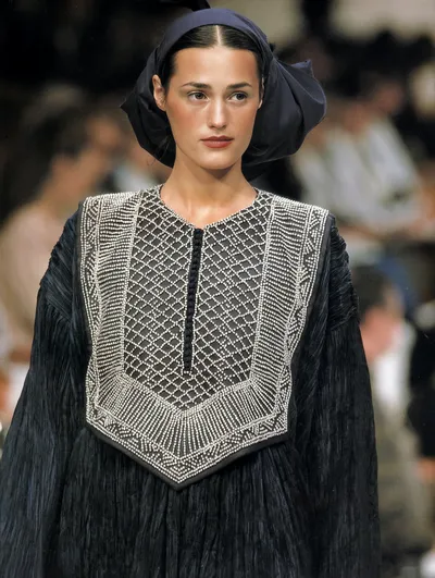 A runway photo of a woman with dark hair pulled back under a large navy blue bow or headscarf, wearing a dark navy pleated dress with an elaborate silver beaded bib-style yoke featuring a diamond lattice pattern.